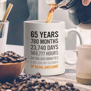 May include: White coffee mug with black text that reads "65 Years, 780 Months, 23,740 Days, 569,777 Hours, 34,186,669 Minutes, 2,051,200,189 Seconds, Of Being Awesome". The mug is being filled with coffee from a glass carafe.