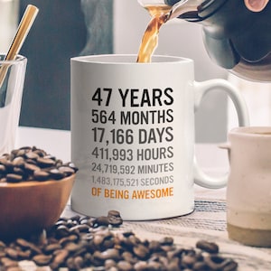 以下が含まれることがあります： 白磁のコーヒーカップに、黒い文字で「47年 564ヶ月 17,166日 411,993時間 24,719,592分 1,483,175,521秒 素晴らしい存在である」と書いてあります。
