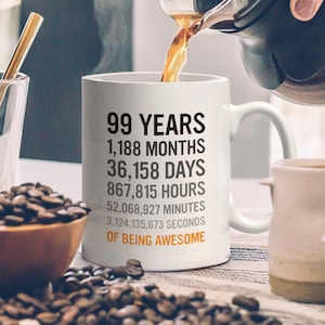May include: White ceramic coffee mug with black text that reads "99 Years 1,188 Months 36,158 Days 867,815 Hours 52,068,927 Minutes 3,124,135,673 Seconds of Being Awesome".