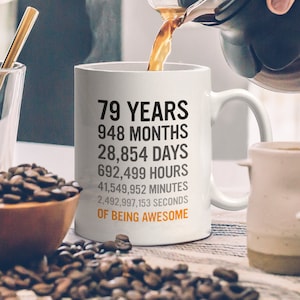 May include: White ceramic coffee mug with black text that reads "79 Years, 948 Months, 28,854 Days, 692,499 Hours, 41,549,952 Minutes, 2,492,997,153 Seconds of Being Awesome".