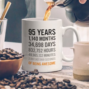 May include: White ceramic coffee mug with black text that reads "95 Years, 1,140 Months, 34,698 Days, 832,752 Hours, 49,965,132 Minutes, 2,997,907,969 Seconds of Being Awesome".