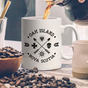 May include: A white ceramic mug with the text "Oak Island Nova Scotia" and a diamond, cross, skull, and ship wheel design is being filled with coffee. Coffee beans and other mugs are also visible.