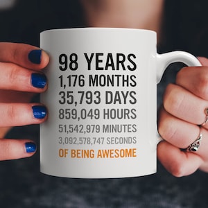 May include: White ceramic mug with black text that reads "98 Years, 1,176 Months, 35,793 Days, 859,049 Hours, 51,542,979 Minutes, 3,092,578,747 Seconds of Being Awesome".