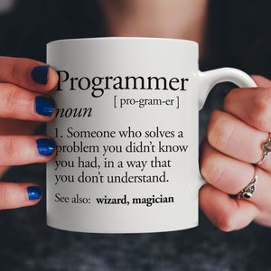 Puede incluir: Taza de cerámica blanca con una definición en blanco y negro de la palabra "Programador" impresa en ella. La definición dice: "Alguien que resuelve un problema que no sabías que tenías, de una manera que no entiendes. Ver también: mago, mago."