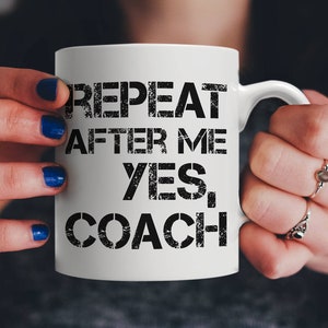 Könnte beinhalten: Weiße Keramiktasse mit schwarzem Text, der "REPEAT AFTER ME YES, COACH" lautet.