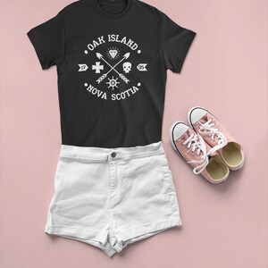 May include: Black t-shirt with a white graphic design that says "Oak Island, Nova Scotia" with a diamond, skull, cross, and compass design. White denim shorts and pink sneakers.