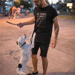 May include: A man wearing a black t-shirt with the text "50 YEARS 600 MONTHS 18,262 DAYS 438,290 HOURS 26,301,438 MINUTES 1,578,086,280 SECONDS OF BEING AWESOME" is playing with a white and black dog.