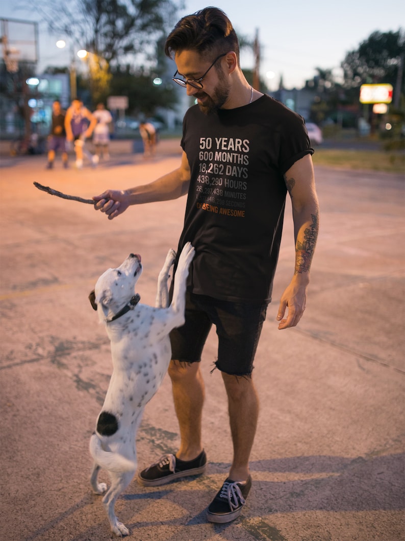 May include: A man wearing a black t-shirt with the text "50 YEARS 600 MONTHS 18,262 DAYS 438,290 HOURS 26,301,438 MINUTES 1,578,086,280 SECONDS OF BEING AWESOME" is playing with a white and black dog.