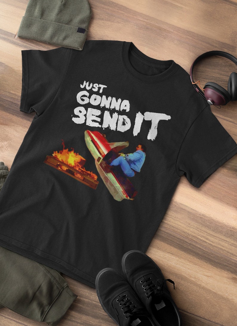 K&ouml;nnte beinhalten: Schwarzes T-Shirt mit einer wei&szlig;en Grafik einer Person auf einem Schneemobil, die &uuml;ber ein Feuer springt. Der Text "JUST GONNA SEND IT" ist in Wei&szlig; &uuml;ber der Grafik geschrieben.