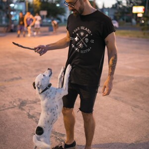 May include: A man wearing a black t-shirt with a white graphic design that says "Oak Island, Nova Scotia" is playing with a white and black dog. The graphic design on the shirt features a compass, a skull and crossbones, and arrows.