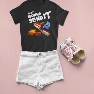 K&ouml;nnte beinhalten: Schwarzes T-Shirt mit wei&szlig;em Text, der "Just Gonna Send It" lautet, und einer Grafik einer Person auf einem Schneemobil. Die Person tr&auml;gt eine rot-blaue Jacke und einen wei&szlig;en Helm. Das Schneemobil ist rot-wei&szlig; und fliegt durch die Luft. Im Hintergrund befindet sich ein Lagerfeuer.