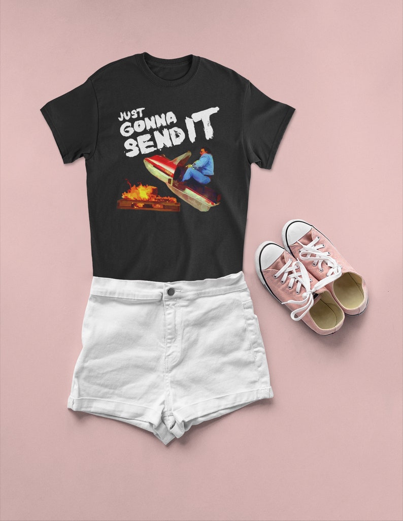 K&ouml;nnte beinhalten: Schwarzes T-Shirt mit wei&szlig;em Text, der "Just Gonna Send It" lautet, und einer Grafik einer Person auf einem Schneemobil. Die Person tr&auml;gt eine rot-blaue Jacke und einen wei&szlig;en Helm. Das Schneemobil ist rot-wei&szlig; und fliegt durch die Luft. Im Hintergrund befindet sich ein Lagerfeuer.