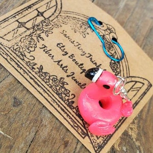 May include: A pink flamingo-shaped charm with a black and white hat, attached to a silver ring. A blue safety pin and a brown card with text and a floral design are also visible. The card reads "Sams Tiny Trinkets Etsy Boutique Fiber Arts Jewels."