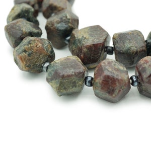 May include: A strand of dark red and green gemstone beads with a faceted, irregular shape. The beads are strung on a black cord.