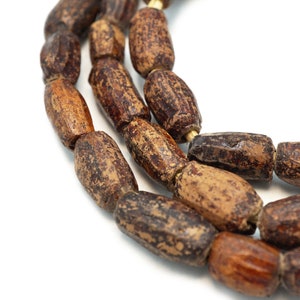 May include: A strand of cylindrical beads with a textured, mottled brown and tan surface. The beads are strung together on a light-colored cord, showing a weathered appearance with variations in color density across each individual bead surface.