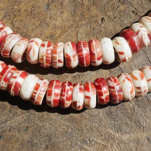 May include: Two strands of beaded necklaces. The beads are a mix of white and red, with a patterned design. The necklaces are displayed on a weathered wooden surface, showcasing the natural colors and textures of the beads.