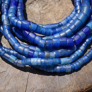 Op de afbeelding: Een verzameling blauwe glazen kralenkettingen. De kralen zijn een mix van vierkante en cilindrische vormen, met verschillende tinten blauw. De kettingen liggen op een verweerd houten oppervlak.
