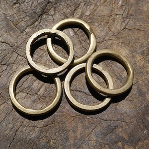 May include: A collection of five brass rings, each with a circular shape and a slightly textured surface. The rings are arranged in a loose pile, with some overlapping. The warm, golden color of the brass contrasts with the dark, wooden background.