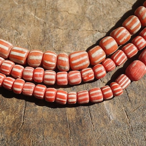 May include: Three strands of orange and white striped beads are displayed. The beads, of varying sizes and shapes, have a handcrafted look. The beads are arranged on a textured, brown surface, giving a rustic feel.