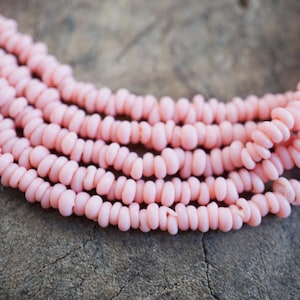 Pode incluir: Close-up de vários fios de contas rosa claro. As contas são pequenas, cilíndricas e enfiadas, criando um efeito em camadas. O fundo é uma superfície texturizada e neutra, proporcionando contraste com a cor rosa suave das contas. Estas contas são adequadas para fazer joias.