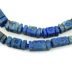 May include: Close-up of two strands of lapis lazuli beads. The beads are a deep blue color with a square shape and carved details. The beads are strung together with small blue beads, creating a necklace or bracelet.