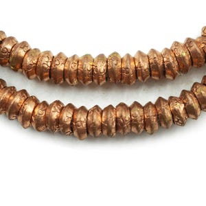 May include: Close-up of two strands of copper-colored beads. The beads are cylindrical with a textured surface and a slightly irregular shape. The beads have a warm, metallic sheen, and are arranged against a white background.