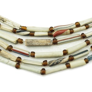 Puede incluir: Primer plano de varios hilos de un collar. El collar presenta cuentas cilíndricas blanquecinas con detalles marrones. Algunas cuentas tienen rayas rojas, azules y negras. Las cuentas están ensartadas con pequeñas cuentas redondas marrones.