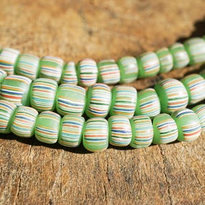 May include: Close-up of three strands of green glass beads. Each bead is cylindrical with horizontal stripes in white, orange, blue, and brown. The beads are strung together, creating a textured and colorful accessory.