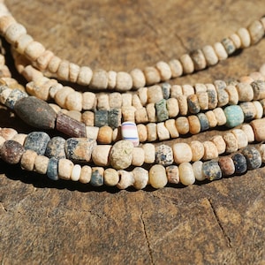 May include: Close-up of several strands of antique beads. The beads are mainly beige, with accents of blue, green, and brown. The beads are strung together, creating a layered effect. The background is a textured, wooden surface.