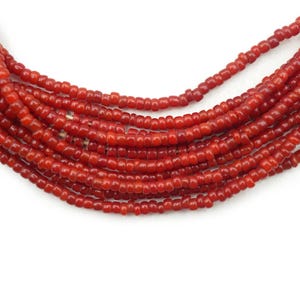 May include: A close-up shot shows multiple strands of small, red beads. The beads are consistent in size and shape, with a slightly rounded, cylindrical form. The strands are arranged in a layered, cascading pattern against a white backdrop, creating a textured, organic appearance.