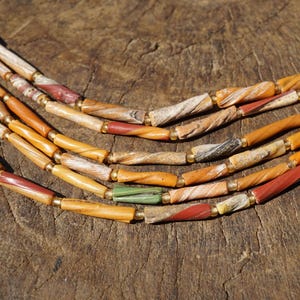 May include: A multi-strand necklace featuring elongated, cylindrical beads in shades of orange, tan, brown, red, and green. The beads have a textured, twisted appearance and are separated by small, metallic-colored spacer beads along the strands.