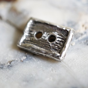 May include: A rectangular, silver-toned button with a textured, wood-grain appearance. The button has two holes for sewing and a slightly weathered look, suggesting a vintage or handcrafted style. The button is approximately 0.75 inches long.