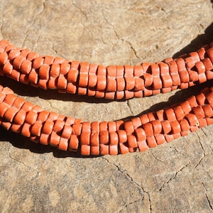 Puede incluir: Primer plano de dos hebras de cuentas de color rojo anaranjado, posiblemente un collar o una pulsera. Las cuentas son rectangulares y están dispuestas en un patrón tejido. El fondo es una superficie marrón texturizada.