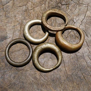 May include: Five antique brass rings of varying sizes and shapes rest on a weathered wooden surface. The rings display a range of patinas, from dark bronze to a lighter, aged gold. Some rings have markings. The arrangement suggests a collection of vintage jewelry.