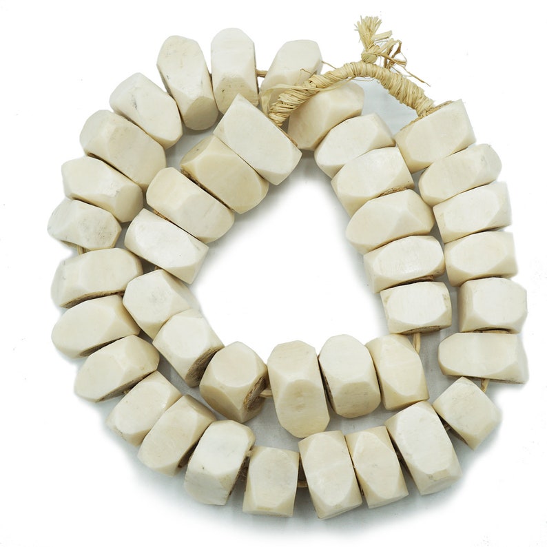 May include: A strand of off-white, geometric beads. Each bead is a rectangular shape with beveled edges. The beads are strung together with a natural fiber cord, creating a necklace or bracelet.