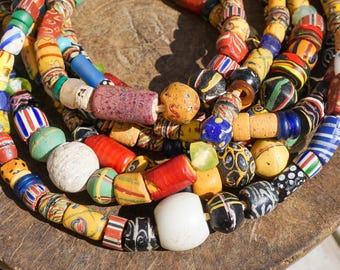 May include: Close-up of several strands of colorful, handcrafted beads. The beads are a mix of shapes, sizes, and colors, including red, yellow, blue, green, and white. Some beads have striped or patterned designs. The beads are arranged on a wooden surface.