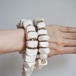 May include: Two off-white, chunky beaded bracelets. The beads are geometric, with a slightly irregular shape. The bracelets are worn on a wrist, showcasing the texture and design of the jewelry. The beads are strung together with a visible string.