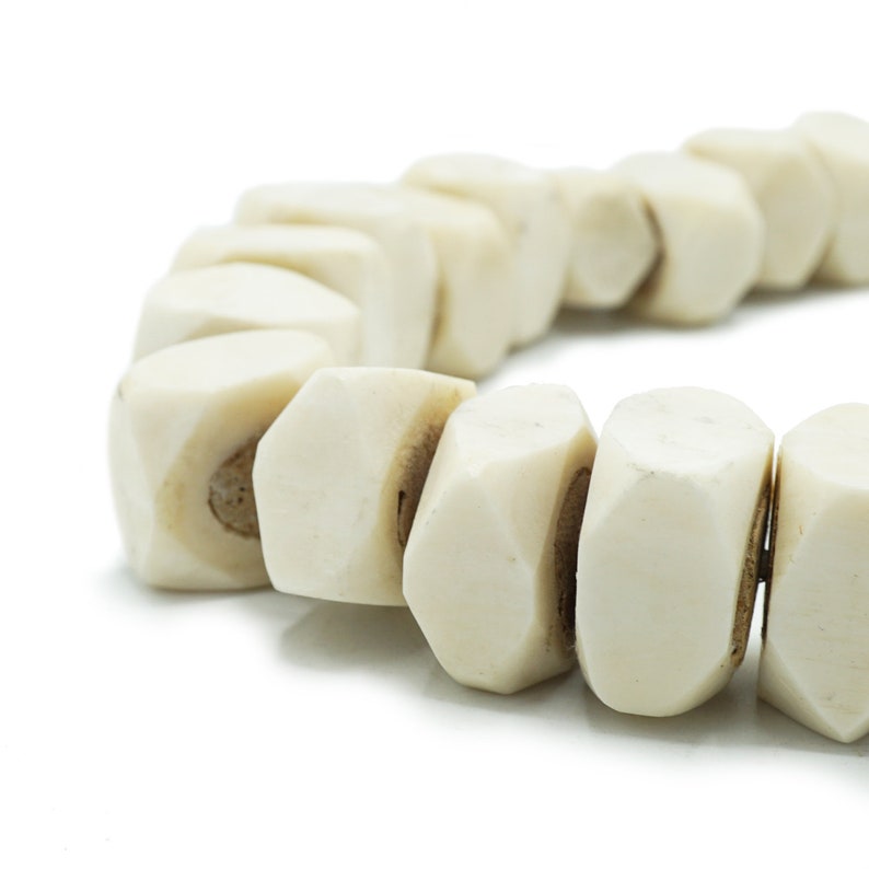 May include: Close-up of a cream-colored beaded bracelet. The beads are geometric, with a faceted design. The bracelet is on a white background. The beads have a natural, organic look.