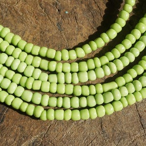May include: Close-up of several strands of light green beads. The beads are small, cylindrical, and evenly spaced, creating a textured surface. The beads are a pale, pastel green color. The background is a rough, brown wooden surface.