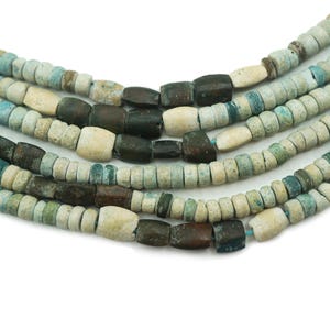 Puede incluir: Primer plano de varios hilos de cuentas antiguas. Las cuentas son una mezcla de colores, incluyendo azul claro, crema y marrón oscuro. Las cuentas son cilíndricas y rectangulares, ensartadas para crear un collar o pulsera.