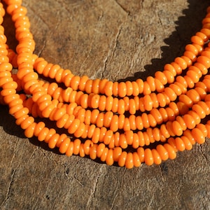 May include: Close-up of several strands of bright orange beads. The beads are small, round, and evenly spaced, creating a textured surface. The beads are resting on a rough, wooden surface.