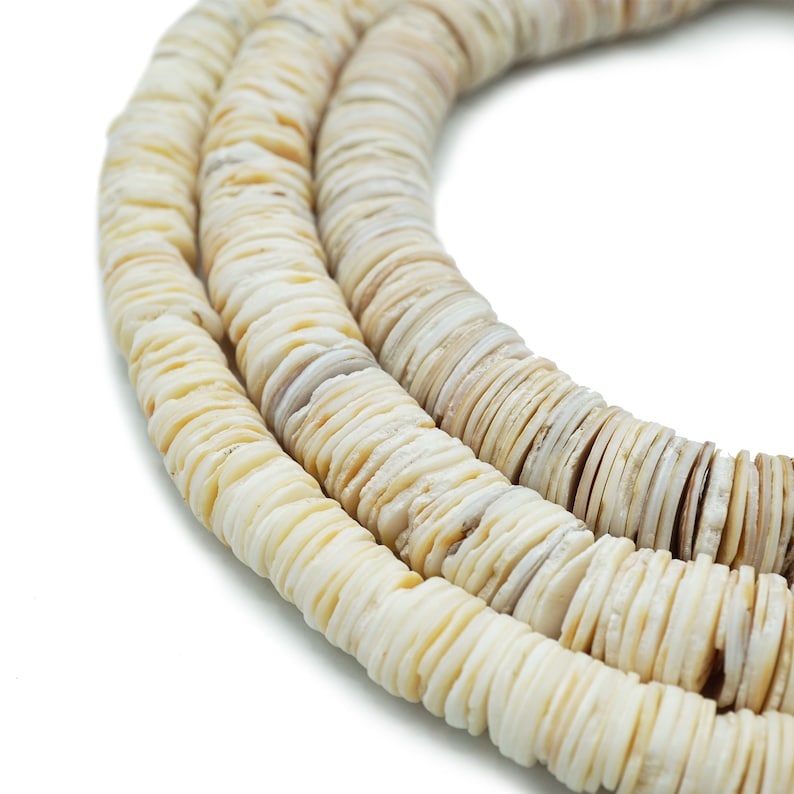 Op de afbeelding: Een streng witte schelp kralen, elke kraal is een platte schijf met een licht onregelmatige vorm. De kralen zijn aan elkaar geregen om een lange ketting te vormen.