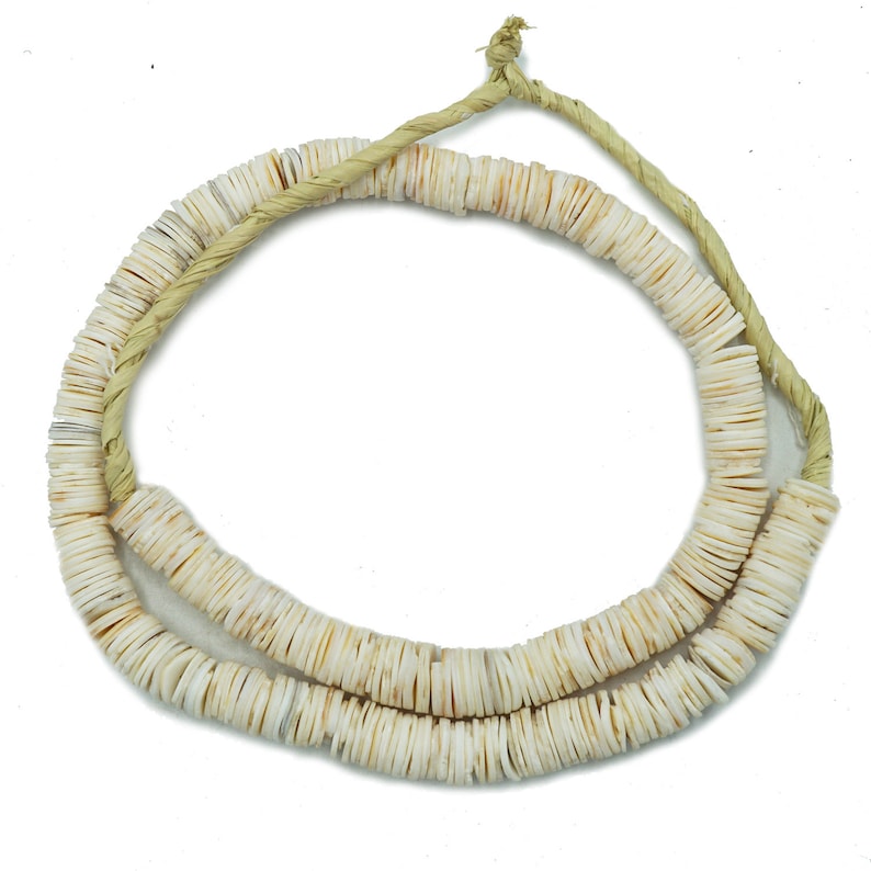 Op de afbeelding: Een witte schelpenketting gemaakt van kleine, ronde, platte schelpen die aan elkaar geregen zijn op een natuurlijke touwtjeskoord.