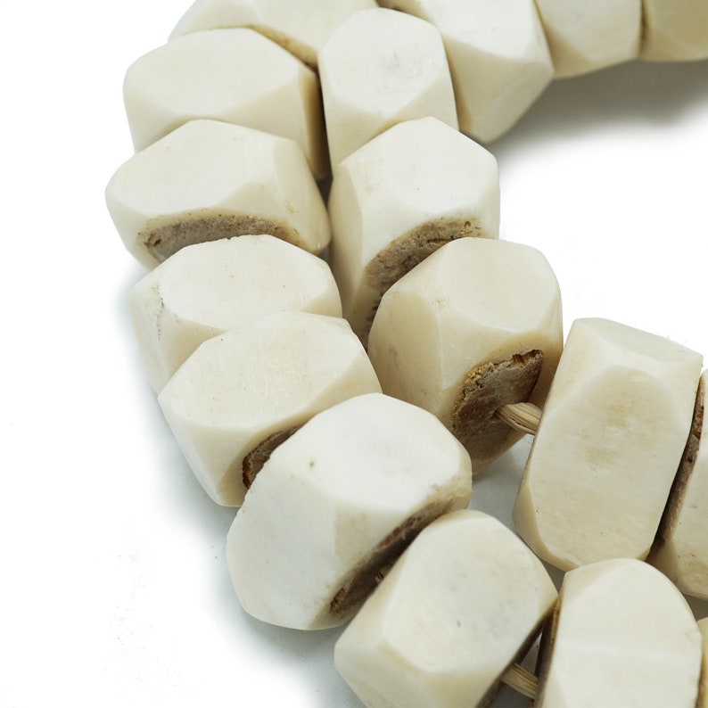 May include: Close-up of a necklace made of off-white, geometric beads. The beads are a light ivory color with a slightly rough texture. Each bead is strung together with a natural-colored cord, creating a unique and earthy aesthetic.