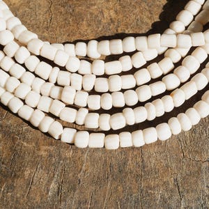 以下が含まれることがあります： クリーム色のビーズネックレスのクローズアップ。 ネックレスは、小さな長方形のビーズの複数のストランドで構成されています。 ビーズは湾曲した線に配置され、木製の質感のある表面に置かれています。 ビーズはマット仕上げです。