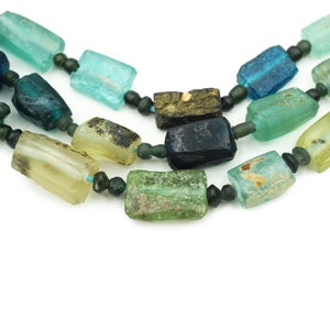 May include: Close-up of three strands of colorful, rectangular glass beads. The beads are various shades of blue, green, and yellow, strung together with smaller, dark green beads. The background is white.