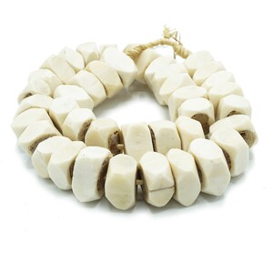 May include: Cream-colored, faceted bead necklace. The beads are strung together with a natural fiber cord. The beads have a geometric shape and a slightly rough texture. The necklace is displayed on a white background.