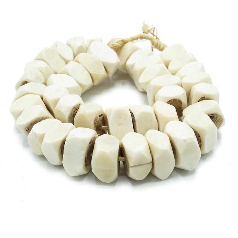 May include: Cream-colored, faceted bead necklace. The beads are strung together with a natural fiber cord. The beads have a geometric shape and a slightly rough texture. The necklace is displayed on a white background.