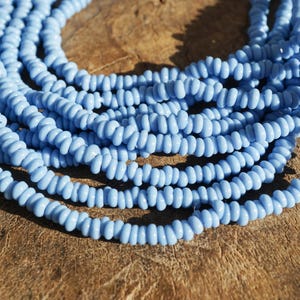 Pode incluir: Close-up de vários fios de contas azul claro. As pequenas contas cilíndricas são enfiadas, criando uma superfície texturizada. As contas estão dispostas sobre uma superfície de madeira castanha, com a luz refletindo nelas.