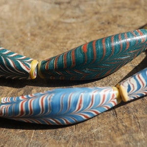 Może przedstawiać: Zbliżenie naszyjnika z dużymi, kolorowymi koralikami. Koraliki mają skomplikowane wzory w kolorze morskim, czerwonym, białym i niebieskim, z żółtymi przekładkami. Naszyjnik jest prezentowany na rustykalnej drewnianej powierzchni.